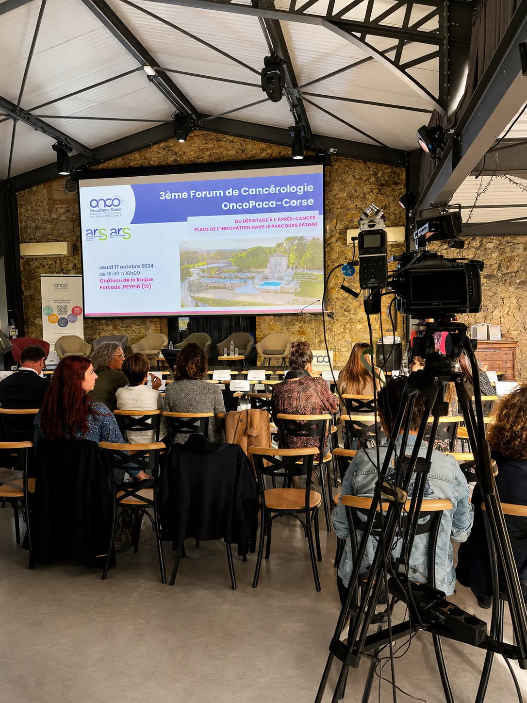 Écran de projection affichant le programme du 3e Forum de Cancérologie OncoPaca-Corse, public assis et caméra de captation au premier plan
