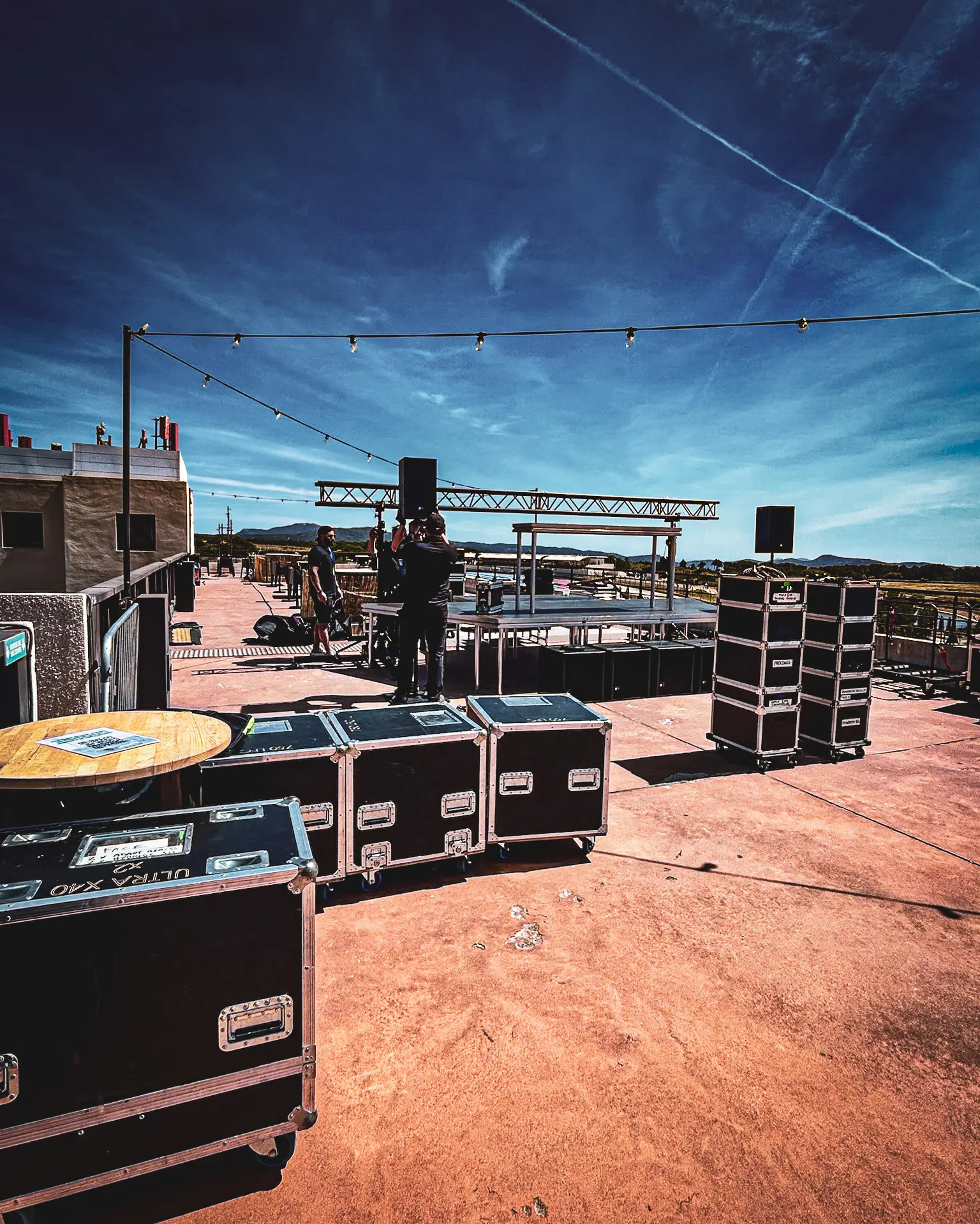 Flight cases et matériel de tournée conditionnés sur le toit d'un lieu événementiel en cours de montage, avec structure d'éclairage et scène en arrière-plan.