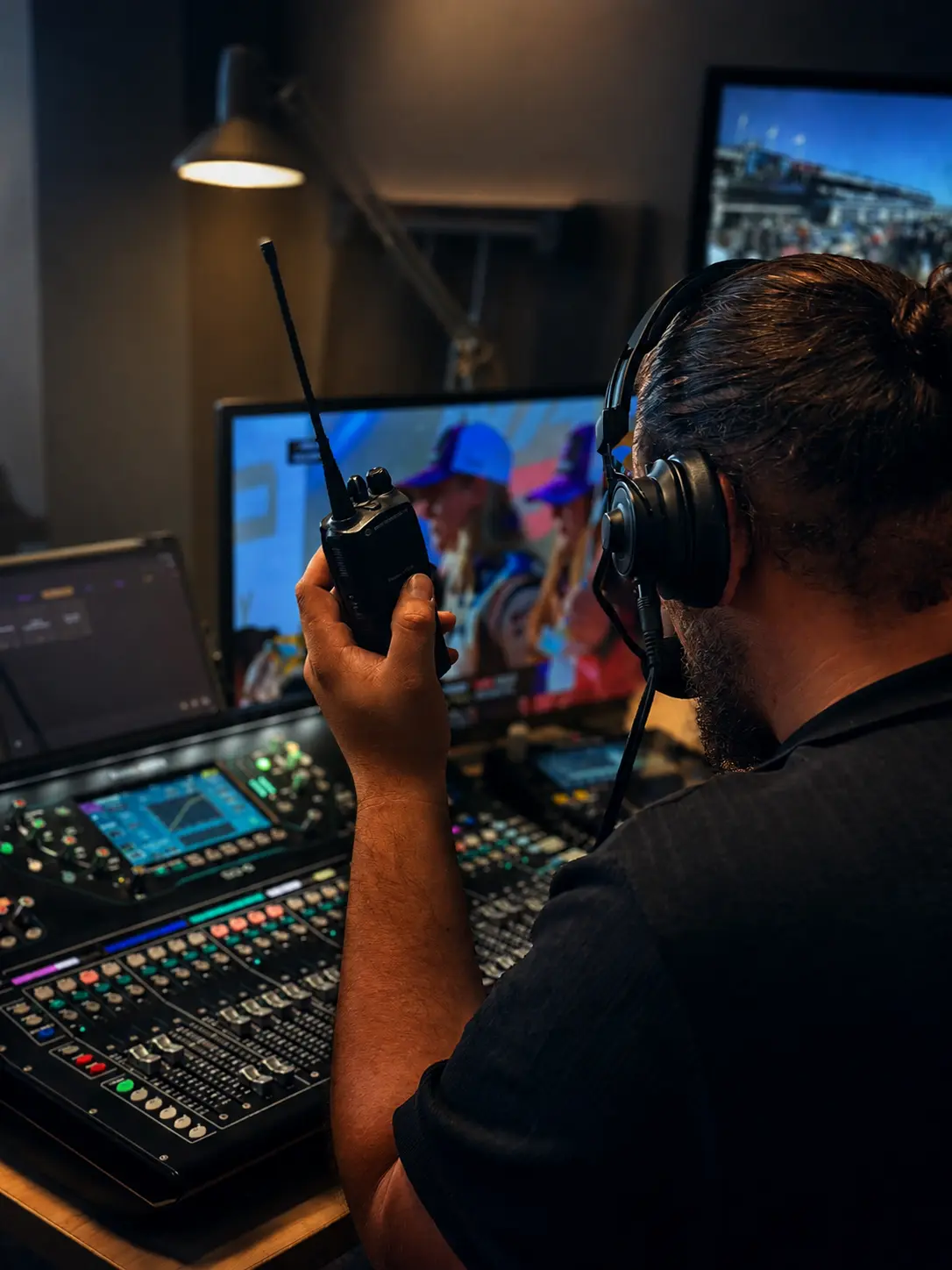 Régisseur de dos, casque de communication et talkie-walkie en main, face à une console de mixage et des écrans de contrôle diffusant un événement sportif.