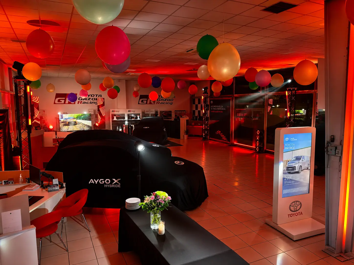 Véhicule Toyota Aygo X Hybride recouvert d'une housse avant reveal, showroom décoré de ballons en lumière rouge