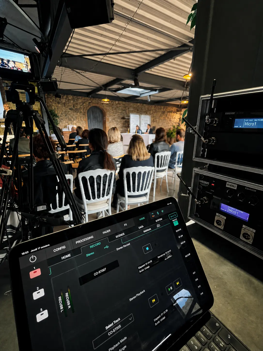 Tablette de régie audio/enregistrement au premier plan, rack de récepteurs micros HF et caméra de captation en situation de conférence